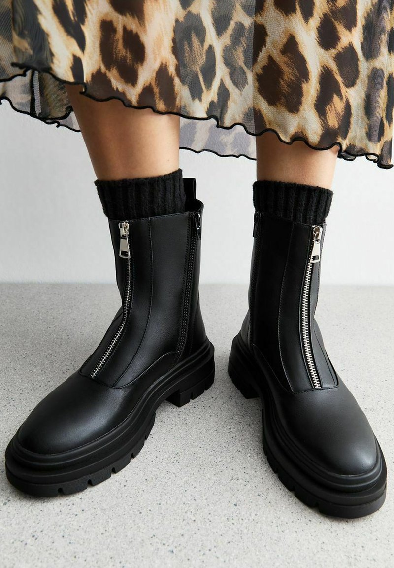 Bottes en cuir noir chunky avec des fermetures éclair à l'avant, portées avec des chaussettes noires côtelées et une jupe imprimée léopard dont l'ourlet est visible au-dessus des chevilles.