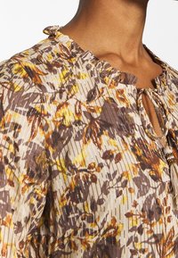 Monivärinen kukallinen pusero, jossa on beige tausta ruskeilla ja keltaisilla kuvioilla, röyhelökaulus ja solmittava yksityiskohta koristehelmellä.