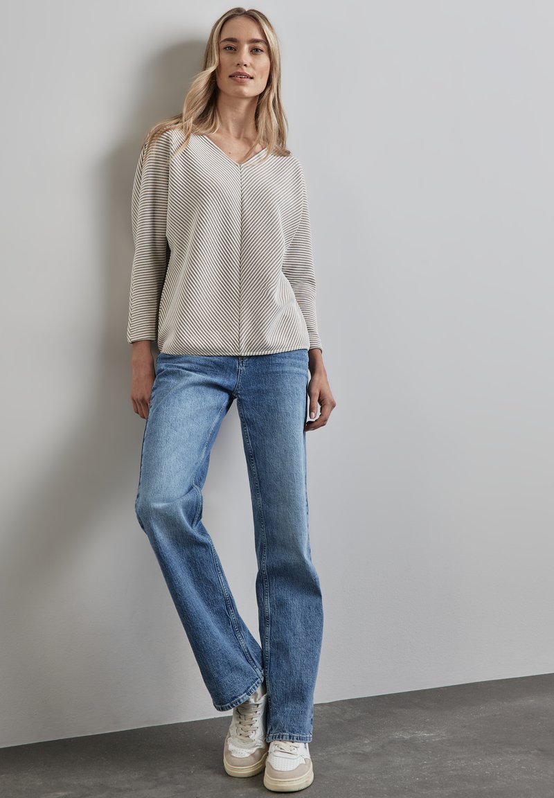 Gestreifter V-Ausschnitt-Pullover in hellbeige und grau, kombiniert mit blauen geraden Jeans und weißen Sneakers. Glattes Material mit einem lockeren Schnitt.