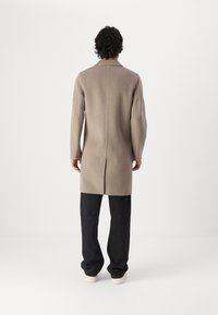 Cappotto di lana beige con taglio dritto, spacco posteriore e maniche lunghe, abbinato a pantaloni neri a gamba larga e scarpe bianche.