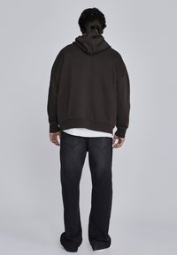 Veľká tmavohnedá mikina s kapucňou na šnúrku, doplnená o čierne džínsy. Hladký povrch s voľným strihom a rebrovanými manžetami.