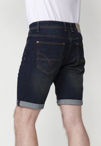 Pantalones cortos de mezclilla en azul oscuro con dobladillos doblados, con costuras naranjas y una etiqueta marrón en la parte trasera, que muestran un diseño clásico.