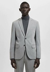 Zwarte man draagt een lichtgrijs colbertjasje en broek met een donkergrijze coltrui, staand tegen een effen lichte achtergrond.