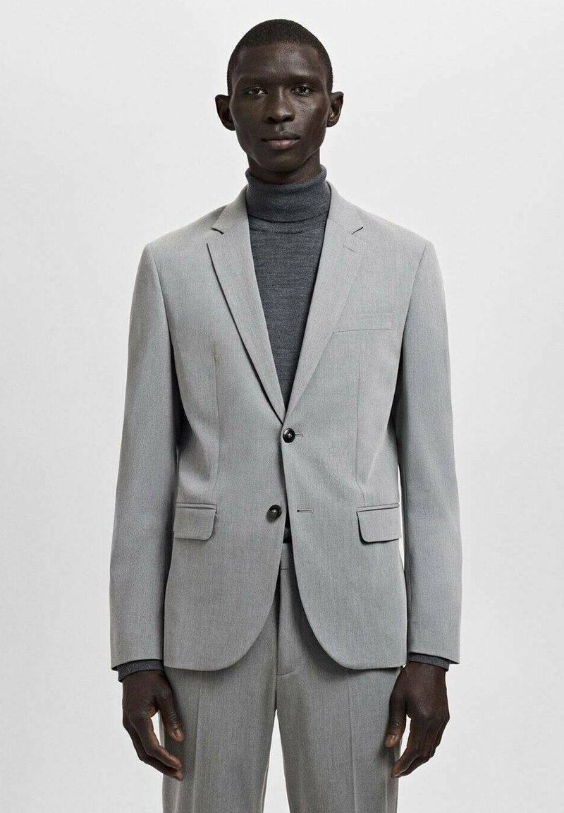 Zwarte man draagt een lichtgrijs colbertjasje en broek met een donkergrijze coltrui, staand tegen een effen lichte achtergrond.