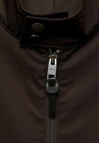 Braune Jacke mit hohem Kragen, ausgestattet mit einem silbernen Reißverschluss und schwarzem Leder-Ziehgriff sowie zwei sichtbaren schwarzen Druckknöpfen. Glatte Textur.