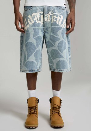 Szorty jeansowe z abstrakcyjnym wzorem i napisem "Ed Hardy", noszone z białymi skarpetkami i jasnobrązowymi sznurowanymi butami na ciemnoskórych nogach.