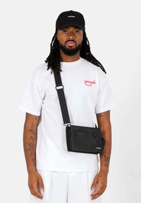 Zwarte schoudertas met een ritsvak, gedragen op een wit T-shirt. Heeft een band en subtiele merknaam. Soepele textuur.