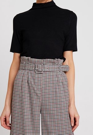 Vrouw die een zwart topje met korte mouwen en een opstaande kraag draagt, gecombineerd met een hoge taille geruite broek en een bijpassende riem.