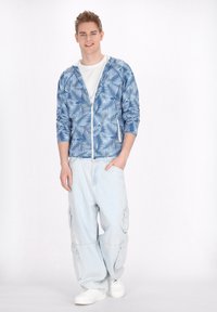 Hanorac cu fermoar de culoare albastru deschis, cu un model de frunze de palmier, asortat cu pantaloni cargo de culoare albastru deschis, cu croială largă, și adidași albi.