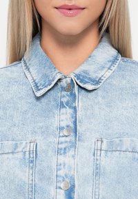 Detailní záběr na dolní část obličeje a krk osoby, která má na sobě lehkou modrou džínovou bundu s potiskem a stříbrnými patentkami a dvojicí náprsních kapes.
