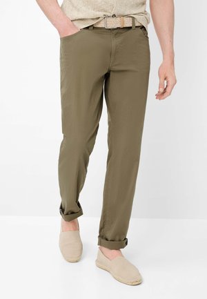 Uomo con pantaloni verde oliva risvoltati, cintura intrecciata beige, scarpe beige slip-on e camicia beige a maniche corte con le mani nelle tasche.
