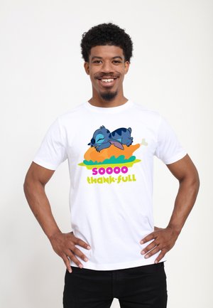 Lächelnder Mann trägt ein weißes T-Shirt mit einer cartoonhaften blauen Figur, die auf einem gebratenen Truthahn schläft, und dem Text „soooo dank-bar.“