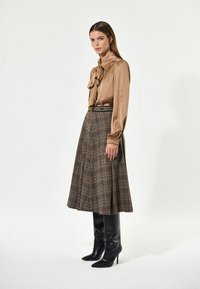 Brun silkebluse med sløjfe og lange ærmer, parret med en plaid midi-nederdel. Sorte knæhøje stilethæle fuldender outfittet.