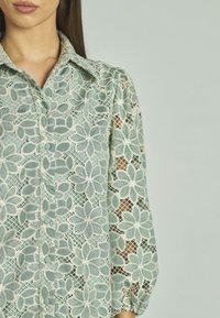 Blusa di pizzo verde chiaro con motivo floreale; presenta un colletto, bottoni e maniche a sbuffo, evidenziando dettagli di design delicati e intricati.