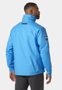 Blauwe waterdichte jas met een hoge kraag, verstelbare manchetten en een logo-patch op de mouw. Soepele textuur en modern ontwerp.
