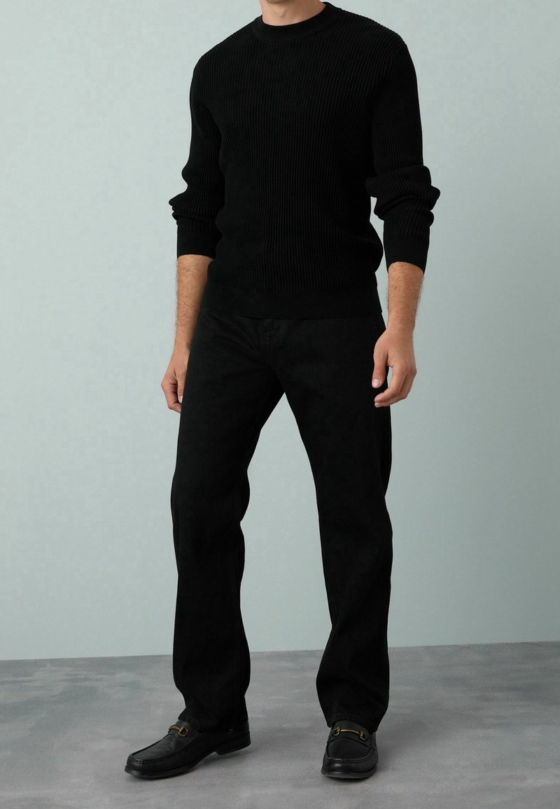Homme debout portant un pull noir côtelé ajusté, un pantalon noir droit et des mocassins noirs sur un sol gris avec un fond gris uni.