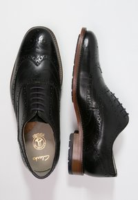 Une paire de chaussures de ville en cuir noir à bout brogue avec des perforations décoratives, une chaussure montrée de dessus, l'autre vue de côté sur fond blanc.