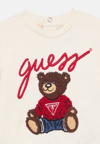 Μπεζ φούτερ με υφασμάτινη κεντημένη καφέ αρκούδα. Η αρκούδα φοράει κόκκινο πουκάμισο με λευκό τριγωνικό λογότυπο και μπλε σορτς. Πάνω αναγράφεται η λέξη «Guess».