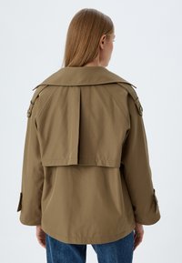 Trench de color caqui con cuello vuelto, puños abotonados y detalles plizados; tejido suave con un ajuste relajado, combinado con unos vaqueros azules.