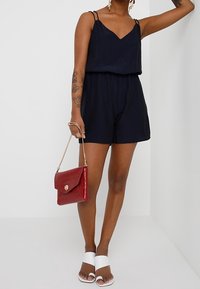 Granatowy kombinezon na ramiączkach, z wcięciem w talii i luźnym krojem. Połączony z białymi klapkami i czerwoną torebką w tłoczony wzór krokodyla.