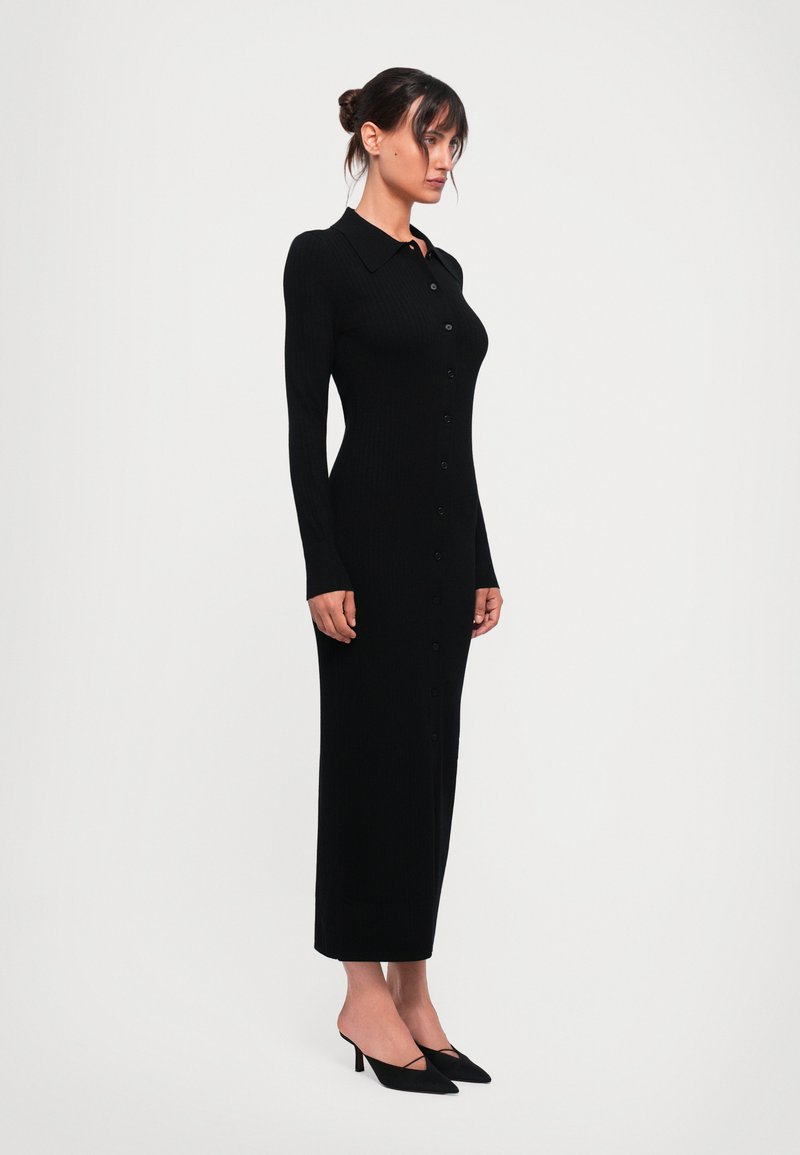 Vestido negro de punto acanalado con cuello, mangas largas y botón completo en la parte delantera. Silueta de lápiz que llega a media pantorrilla, combinado con mule negros de tacón.