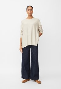 Beige Strickpullover mit entspanntem Schnitt und Rundhalsausschnitt, kombiniert mit dunkelblauen, weit geschnittenen Jeans und braunen Slippers.