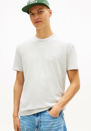 Jonge man die een lichtgrijs T-shirt, een blauwe spijkerbroek met zijn rechterhand in de zak, en een groene baseballpet met een wit logo aan de voorkant draagt.