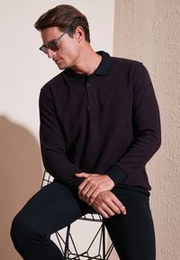 Tmavofialová polokošeľa s dlhým rukávom a čiernym golierom a manžetami, skombinovaná s priliehavými čiernymi nohavicami. Model sedí na drôtenej stoličke.