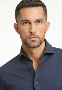 Mörkblå knapp-up skjorta med en texturerad tyg, påfallande krage och två knappar vid knappslånen. Tätt sittande modern design.