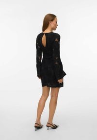 Femme de profil portant une mini-robe noire en dentelle à manches longues avec une découpe en forme de goutte d'eau dans le haut du dos, associée à des escarpins noirs à brides arrière.