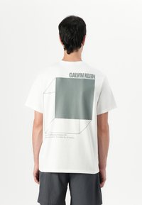 White cotton t-shirt featuring a graphic design of overlapping squares in green on the back, with "CALVIN KLEIN" printed above it.