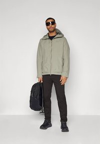 Svetlo siva jakna z zadrgo in kapuco, črne hlače in temni supergi. Drži črn nahrbtnik, nosi velike sončna očala. Enostavno ozadje.