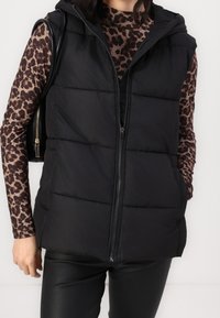 Svart pufferväst med huva, med dragkedjestängning, sidofickor och en quiltad design. Bärs över en långärmad topp med leopardtryck.