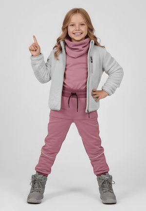 Młoda dziewczyna w różowym stroju na zewnątrz i szarej kurtce, stojąca z rozstawionymi nogami, uśmiechająca się, wskazująca palcem w górę prawą ręką.