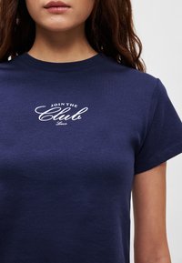 Tumši zils kokvilnas T-krekls ar īsām piedurknēm, uz kura priekšpuses centrā ir balti vārdi “JOIN THE Club” rakstīti kursīvā fontā.