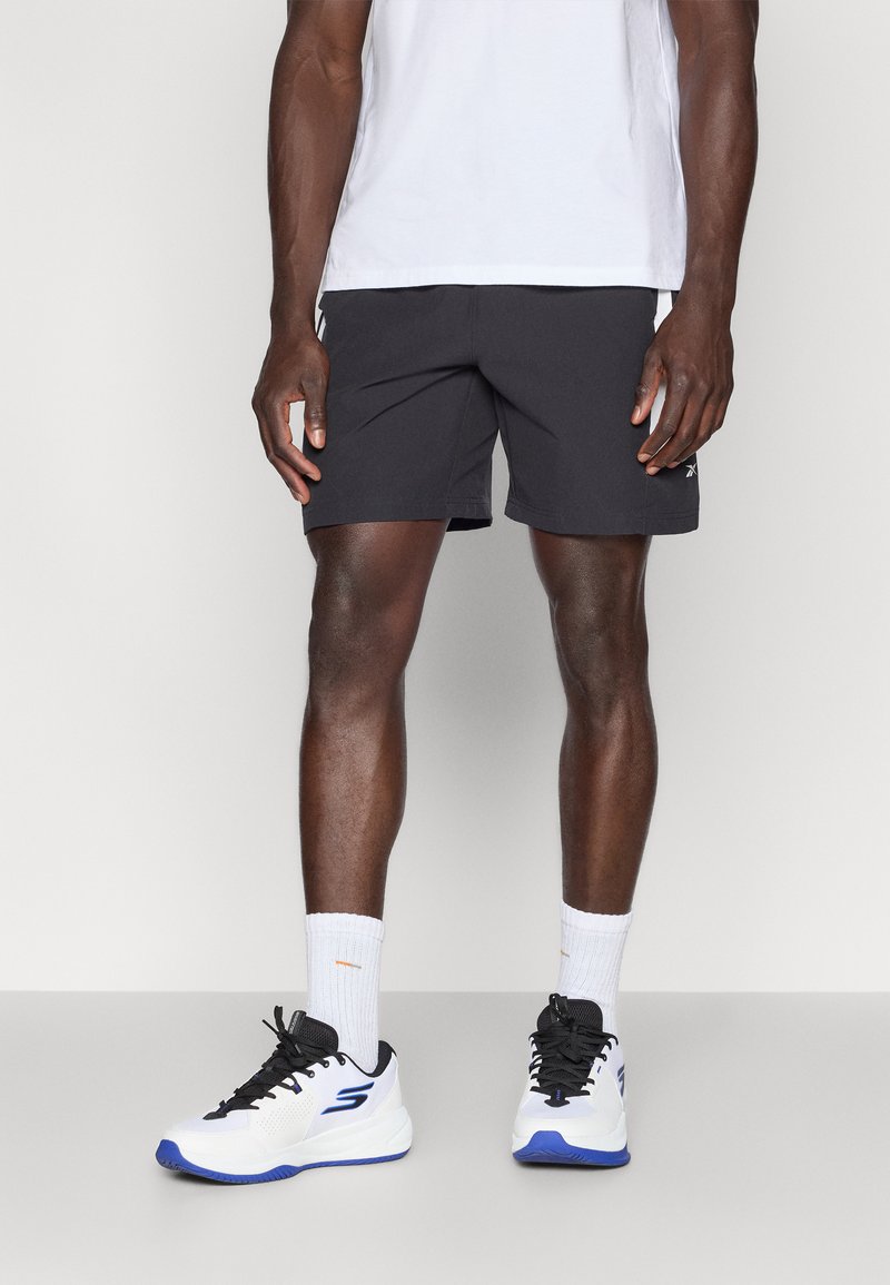 Černé sportovní šortky vyrobené z lehkého materiálu, které jsou kombinovány s bílými teniskami s černými a modrými akcenty. Outfit doplňují bílé kotníkové ponožky.