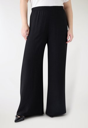 Pantaloni neri a gamba larga con elastico in vita, indossati con scarpe nere a punta e un top bianco, mostrati dalla vita ai piedi.