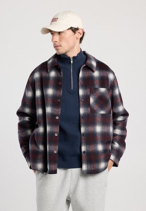 Red and navy check flannel shirt with a button-up front, single chest pocket, worn over a navy mock-neck top, paired with gray sweatpants.