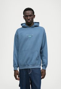 Felpa in cotone blu con tasca frontale e logo sul petto. Il tessuto ha una texture liscia e una vestibilità comoda.