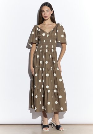 Jonge vrouw in een bruin midi-jurk met witte polka dots, pofmouwen en een geplooide taille, met zwarte open-toe hoge hak sandalen.