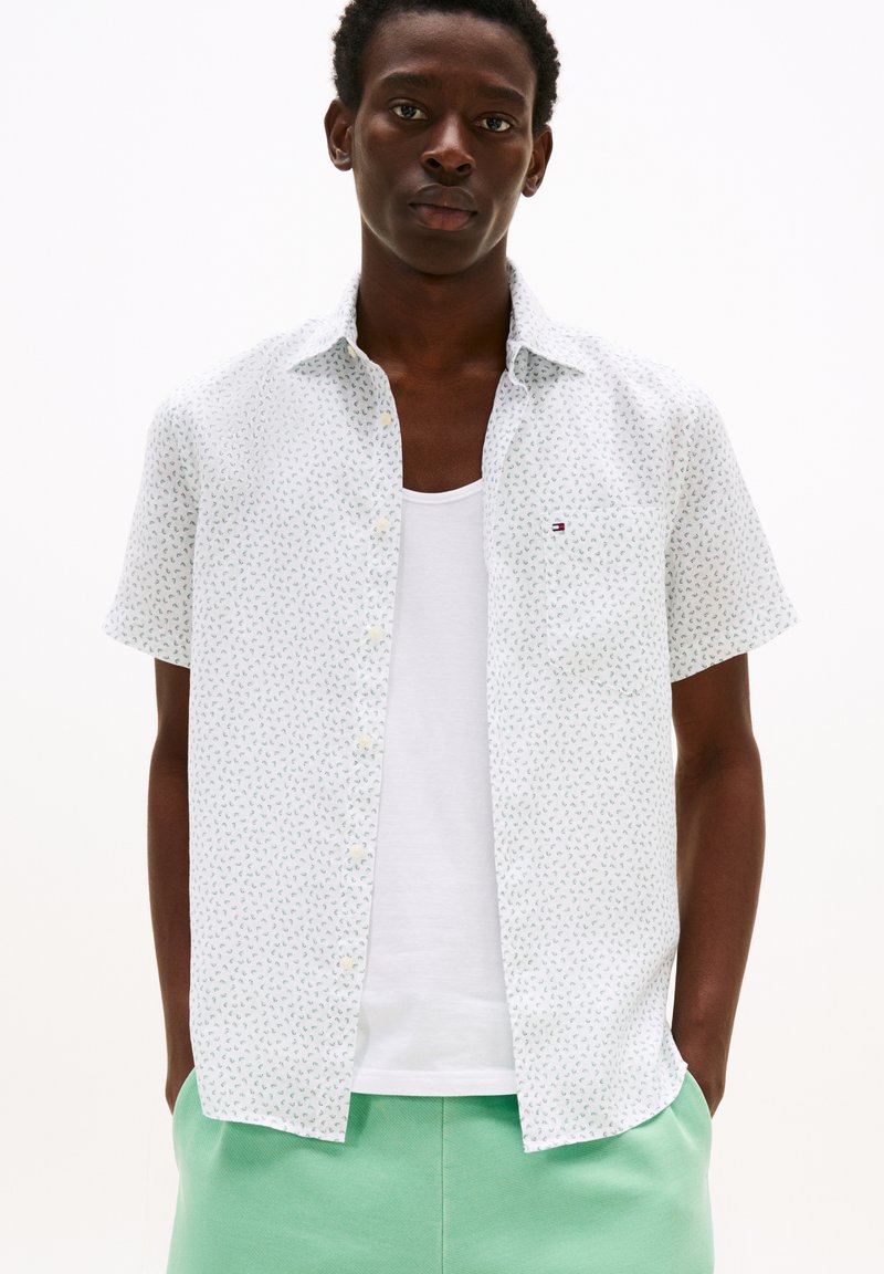 Chemise à manches courtes blanche avec un motif floral vert et bleu. Présente un col boutonné et une petite poche poitrine.