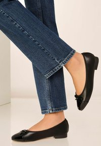 Schwarze Leder-Ballerinas mit runder Zehenpartie, Schleifenakzent an der Vorderseite und niedrigem Absatz, kombiniert mit dunkelblauen Jeans.