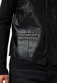 Schwarze Lederweste mit strukturiertem Finish, fünf Knöpfen und einer einzelnen unteren Tasche. Getragen über einem schwarzen Langarmshirt.