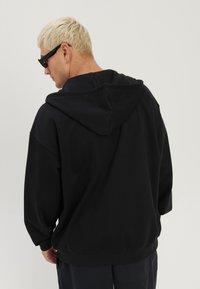 Blondyn ubrany w czarne okulary przeciwsłoneczne i oversize'ową czarną bluzę z kapturem, stojący tyłem na jednolitym jasnym tle.