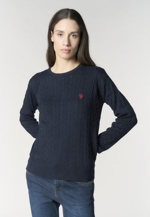 Giovane donna con capelli castani lunghi indossa un maglione blu navy a trecce con logo rosso e jeans blu, in piedi con le mani dietro la schiena.