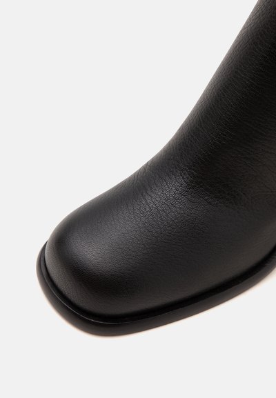Gros plan de la partie avant d'une botte en cuir noir avec un bout arrondi et une surface lisse texturée sur fond blanc.
