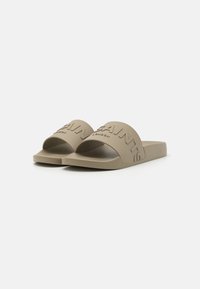 Sandales à glisser beiges avec de larges brides embossées avec "SAINT" et "LONDON". Fabriquées dans un matériau doux et texturé, avec une semelle plate et un design à bout ouvert.