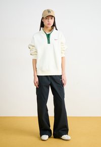 Balts kažoks ar zaļo rāvējslēdzēju, purpura logo; brīvas melnas cargo bikses; baltas sporta kedas; bēša cepure ar logo, gluda tekstūra, brīvā piestāvē.