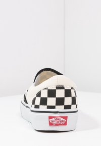 Chaussure à enfiler en toile crème avec un motif à carreaux noirs sur les côtés. Logo Vans rouge sur le talon et semelle en caoutchouc noire.