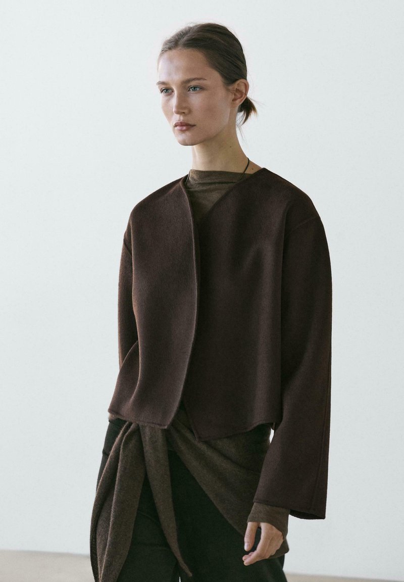 Brown, cropped, open-front jacket with wide sleeves, made of soft fabric. Worn over a dark brown long-sleeve top, creating layered texture.